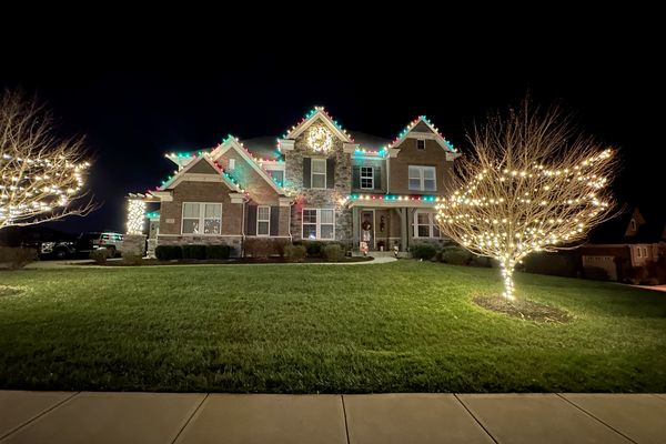 Stone and brick home with multicolor Christmas lights on the roofline and a front-yard tree wrapped in warm white lights